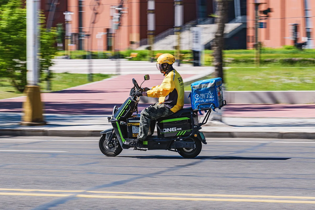 黄色いユニフォームで饿了么(Ele.me)の配達ボックスを積んだ電動バイクが街を走る様子